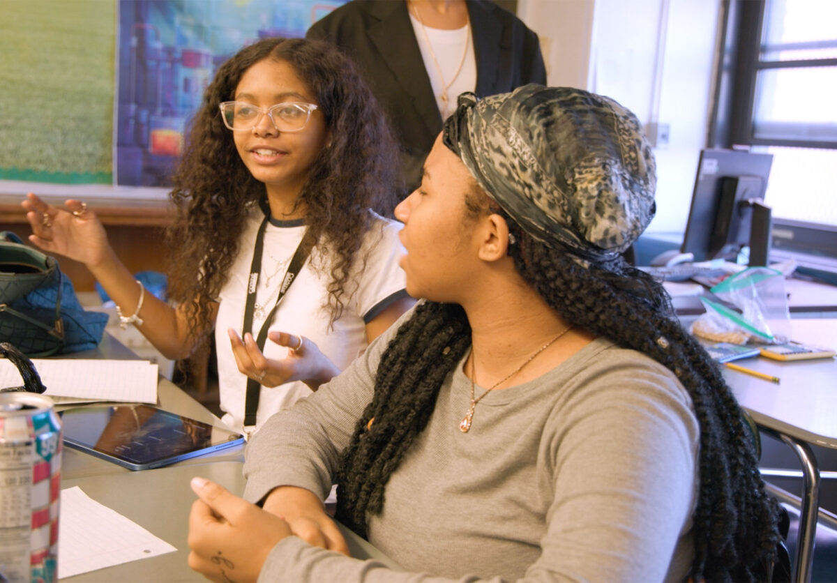 Young women having an animated discussion in a media production class - they are at desks with an ipad and a teacher partly visible behind them.